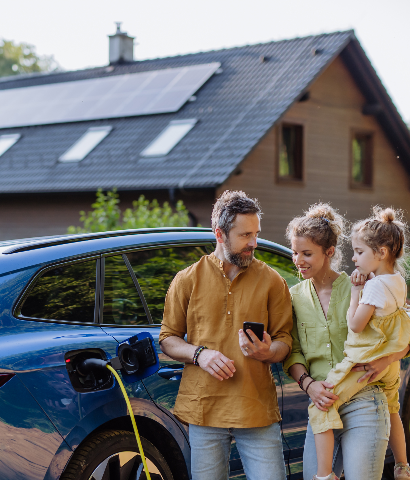 Family of three standing beside a blue electric car that is charging, with a house featuring solar panels in the background.